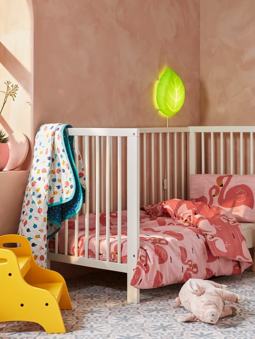 A GULLIVER cot covered in RÖRANDE bedlinen in pink with a RÖRANDE blanket hanging on the side and TROGEN step stool nearby.