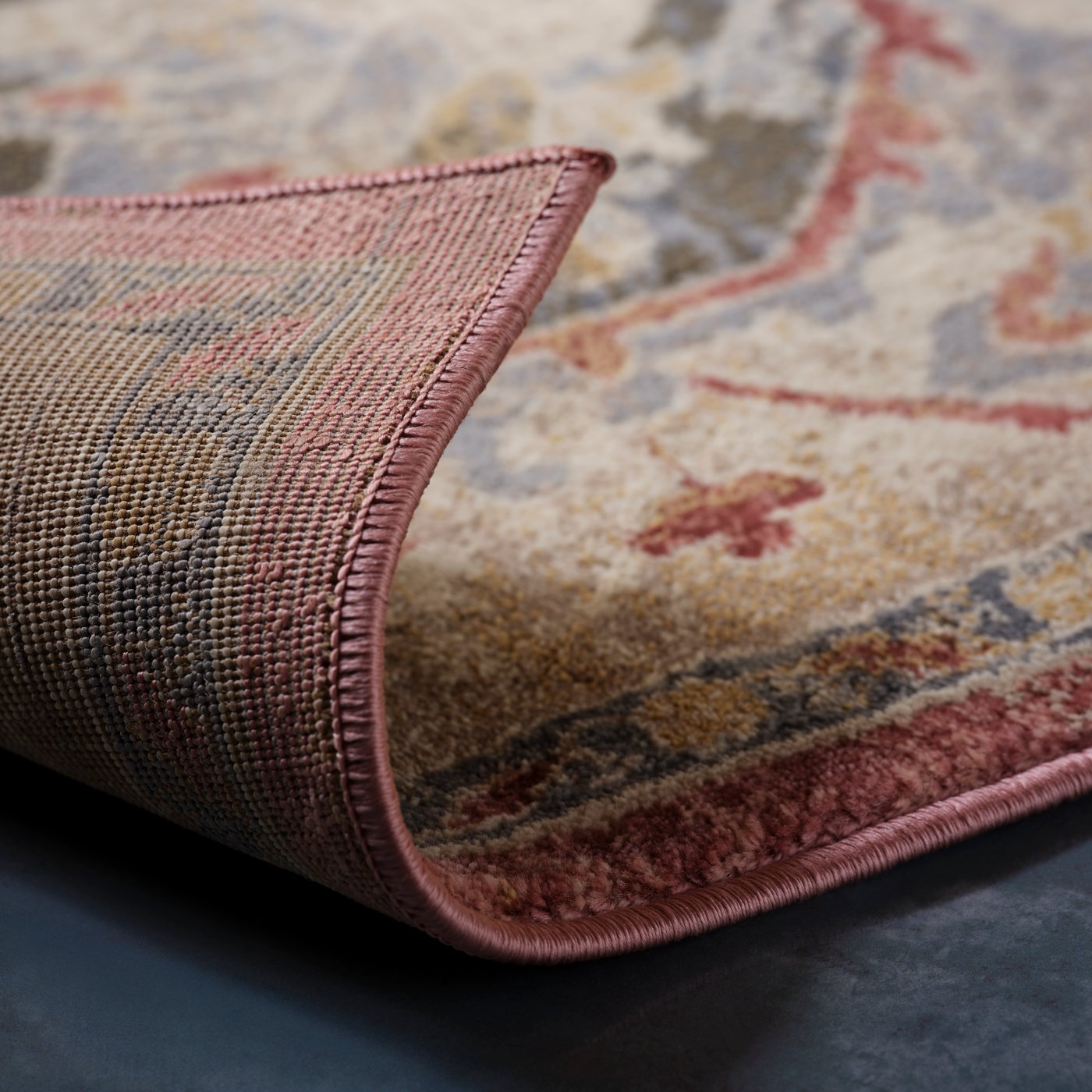 Ett uppvikt hörn av en rosabeige JÄRNVÄG matta med ornamentmönster och låg lugg, som visar undersidans sömmar och detaljer.