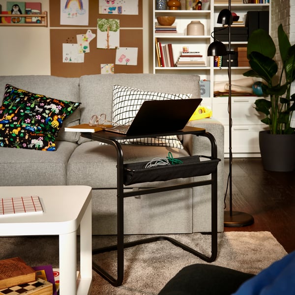 Salon avec table d’appoint OLSERÖD en anthracite/gris foncé sur laquelle est posé un ordinateur portable, à proximité d’un canapé gris garni de coussins à motifs.
