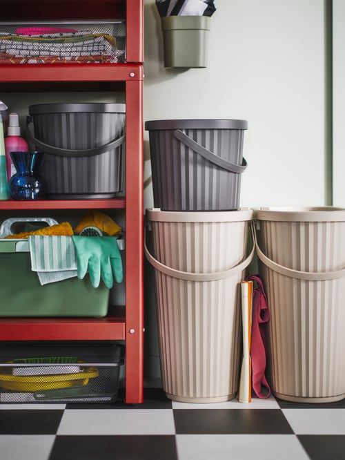 A DAMMÄNG bin with lid is placed alongside other bins next to a storage combination unit filled with cleaning supplies.