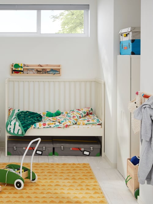 Bedroom section with a cot with a 3-piece bedlinen set, a rug and toddle truck on the floor, and a wardrobe to the side.