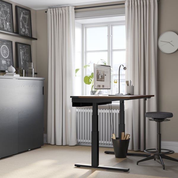 An electric walnut veneer/black MITTZON desk sit/stand in the home office next to a window with curtains hanging.