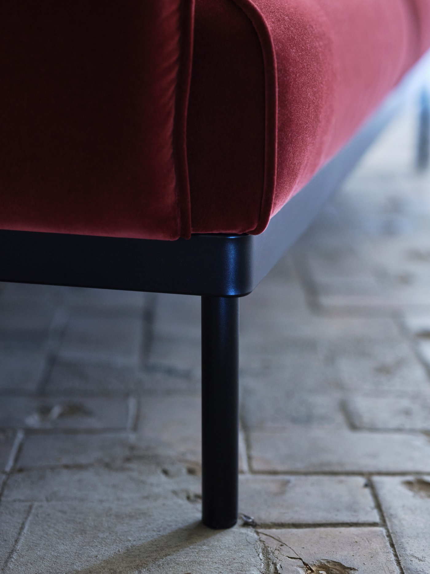 Close-up of the steel leg of a red-brown ÄPPLARYD two-seat sofa on a stone-paved floor.