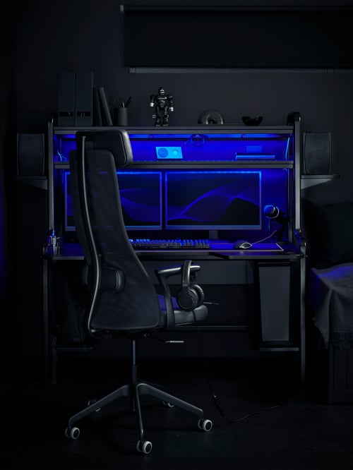 A black FREDDE desk has blue lighting and two computer screens inside. It is in a black room by a black swivel chair.