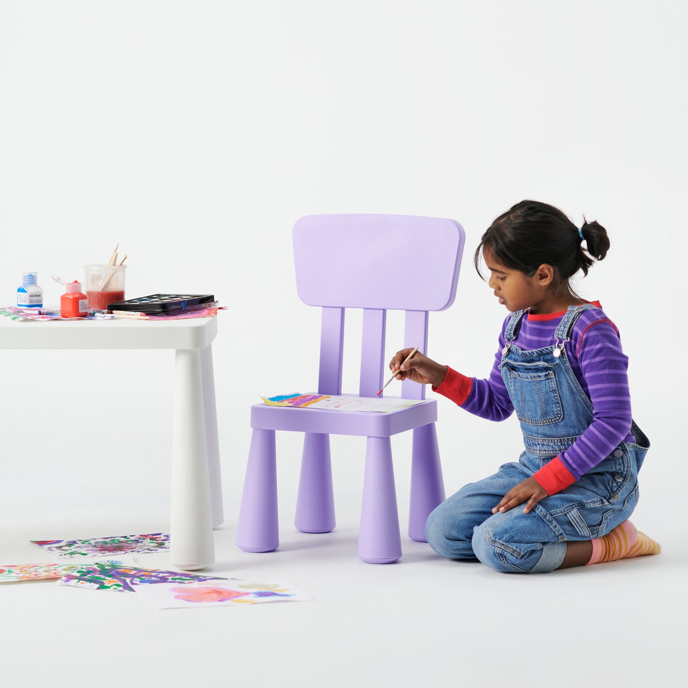 A child painting while using a lilac MAMMUT chair as a tabletop in a clean white studio setting.