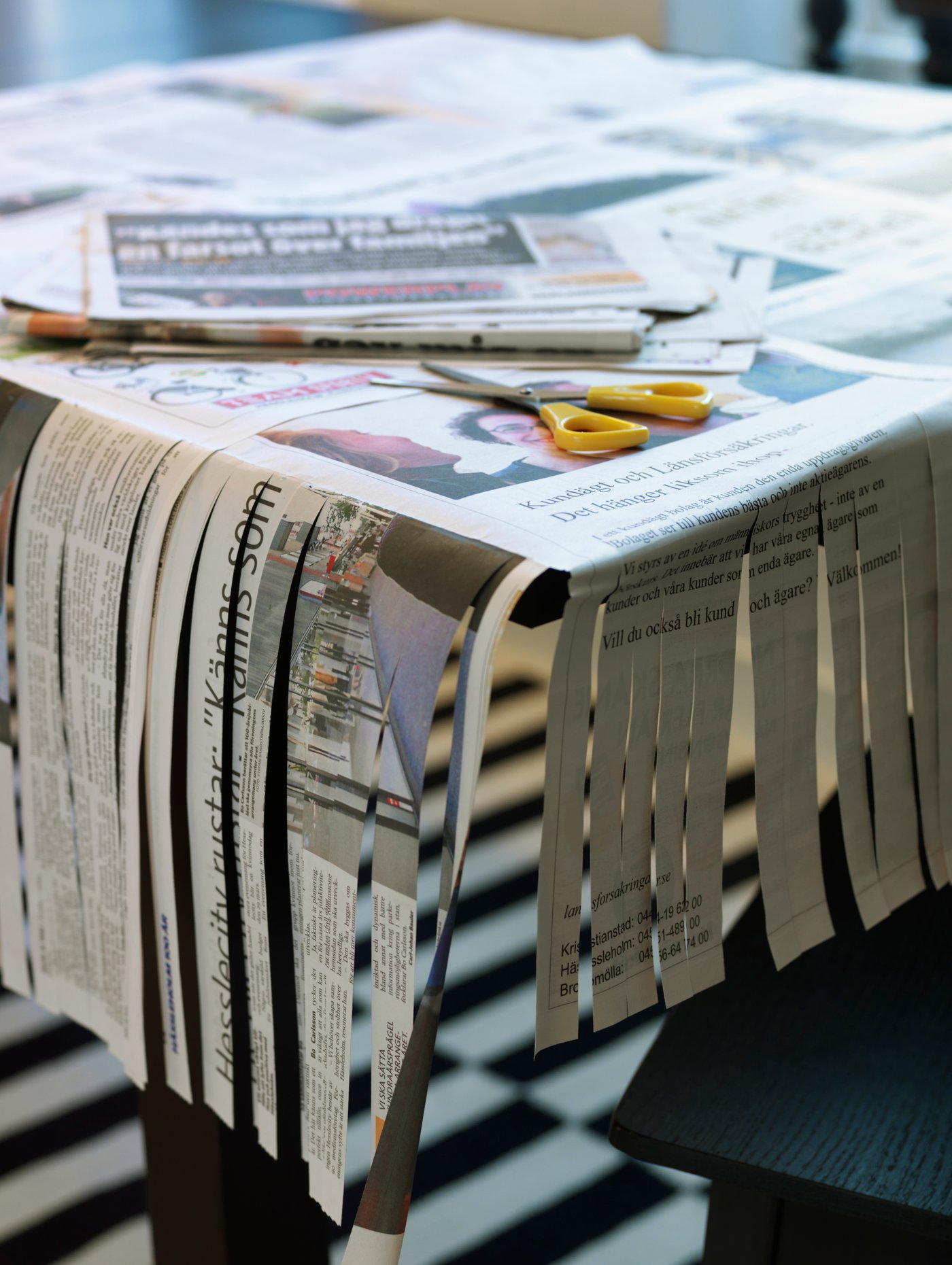 Tisch mit Zeitungen bedeckt, teils in Streifen geschnitten. Gelbe Schere liegt auf dem Tisch. Schachbrettmuster am Boden. "Hasslarp" auf Zeitung.
