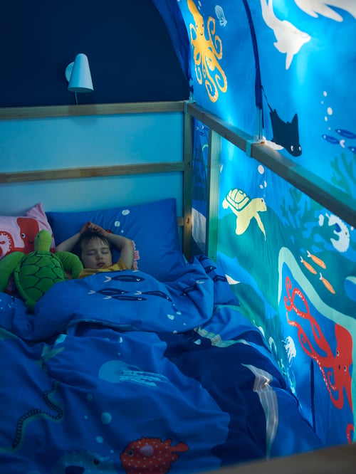 A bed tent surrounds a child sleeping in a reversible bed with a blue duvet cover and a green soft toy.