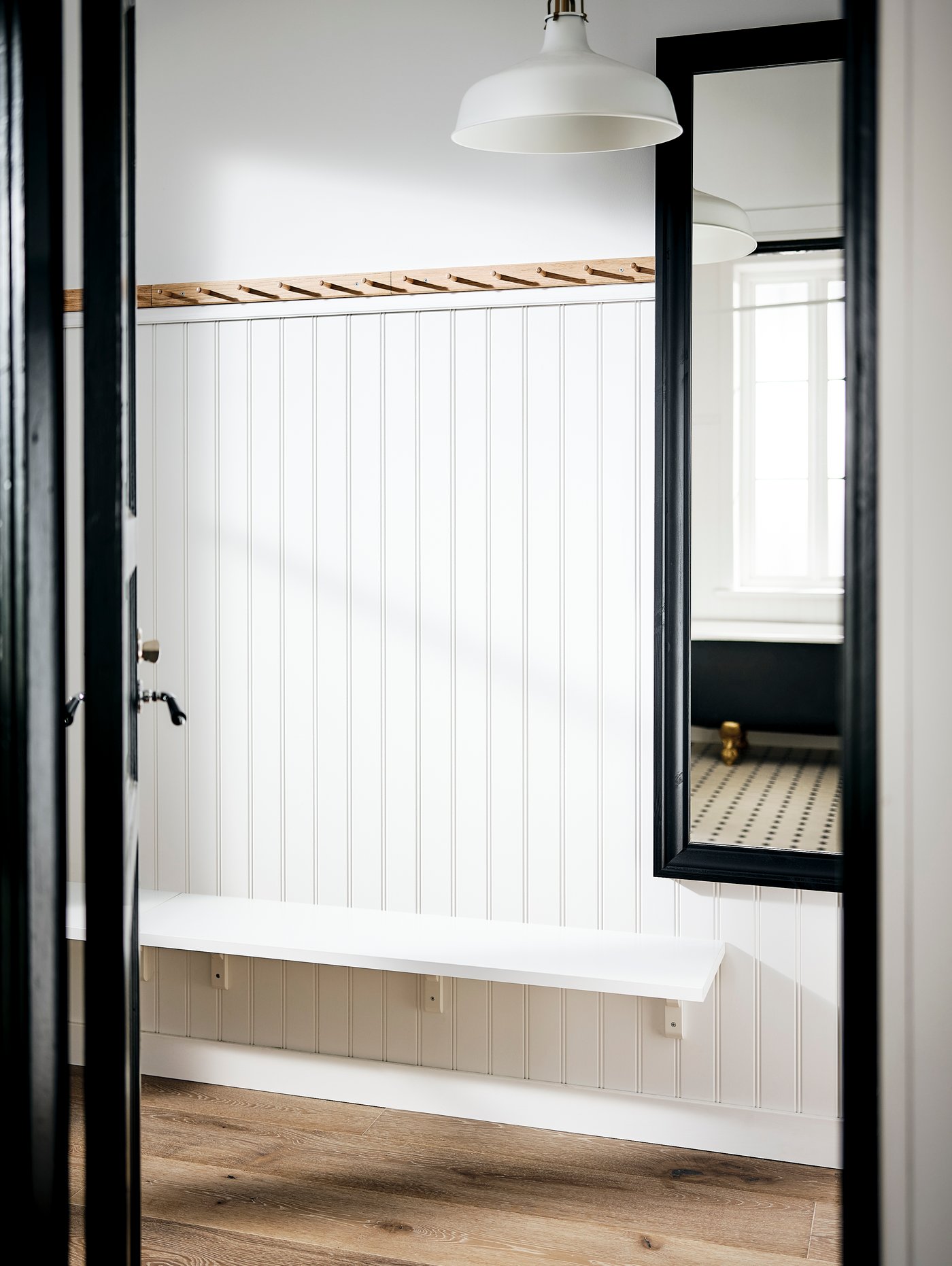 Ett ljust rum med vit pärlspontvägg, lång vit bänk, stor svart ramad spegel och en vit pendellampa hängande ovanför.