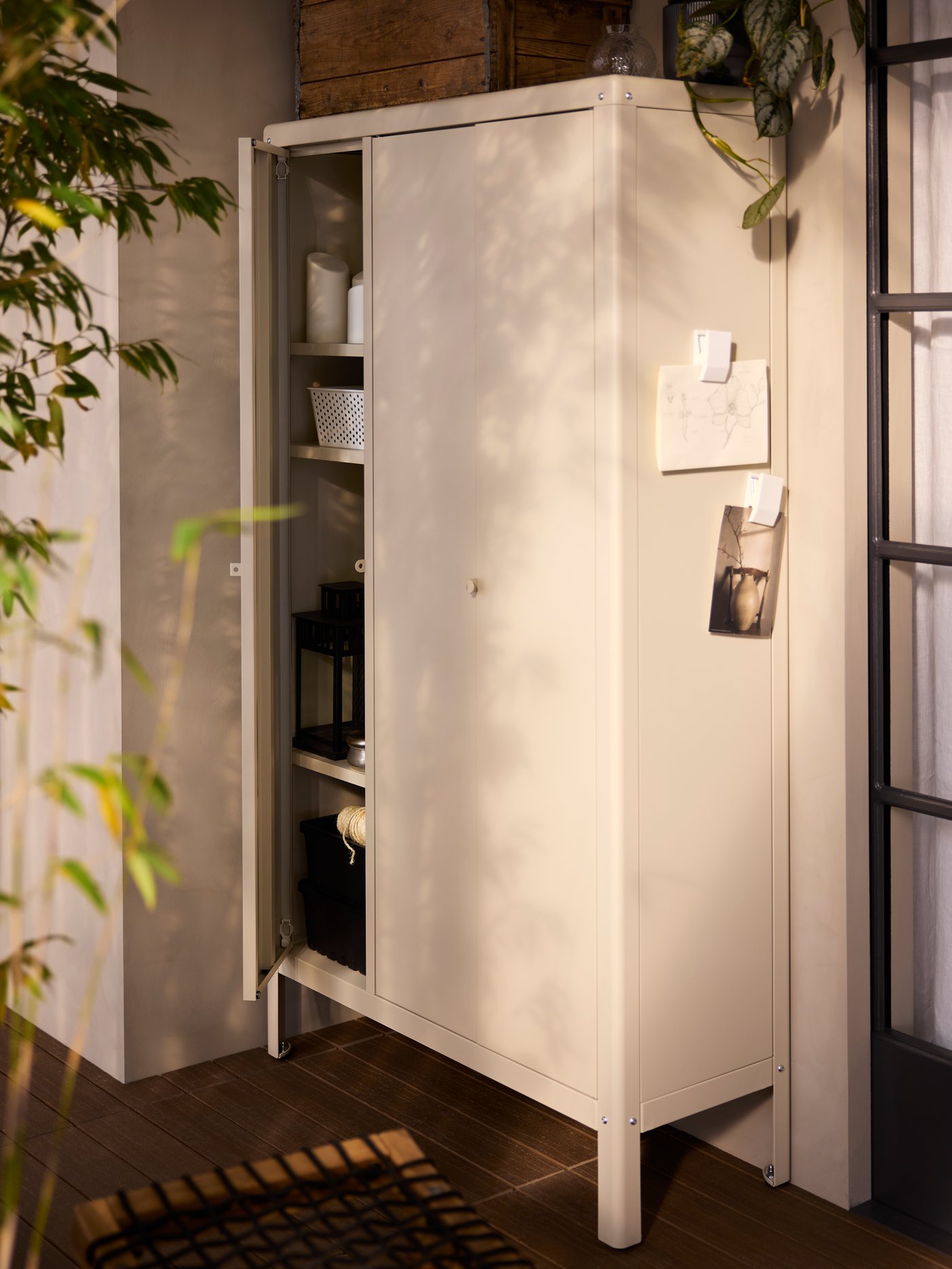 A beige, coated metal KOLBJÖRN in/outdoor cabinet has one of its 3 doors open, showing baskets and lighting on its shelves.