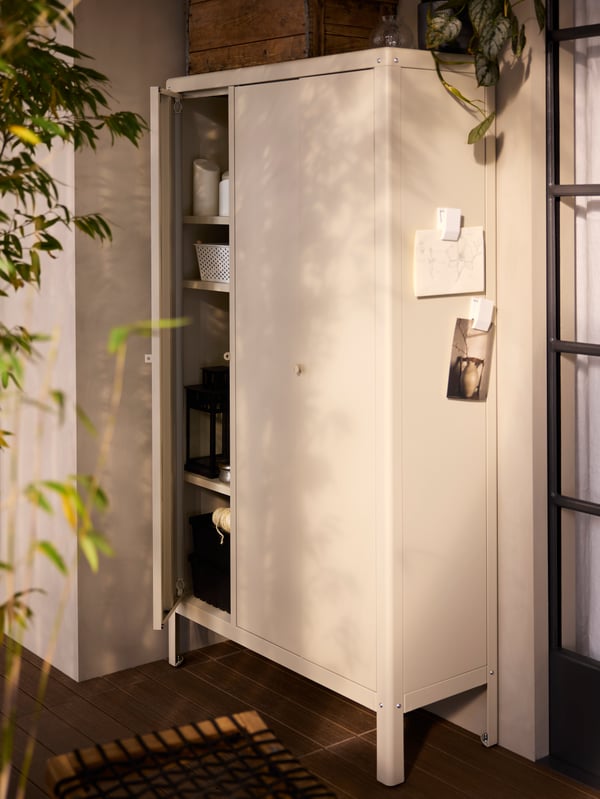 A beige, coated metal KOLBJÖRN in/outdoor cabinet has one of its three doors open, showing baskets and lighting on its shelves.