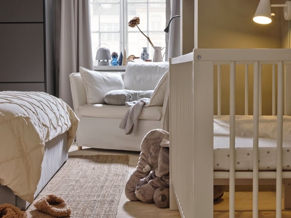 A white BACKSÄLEN 1.5 seat armchair in front of a window with a white GULLIVER cot and a soft toy elephant in front of it.