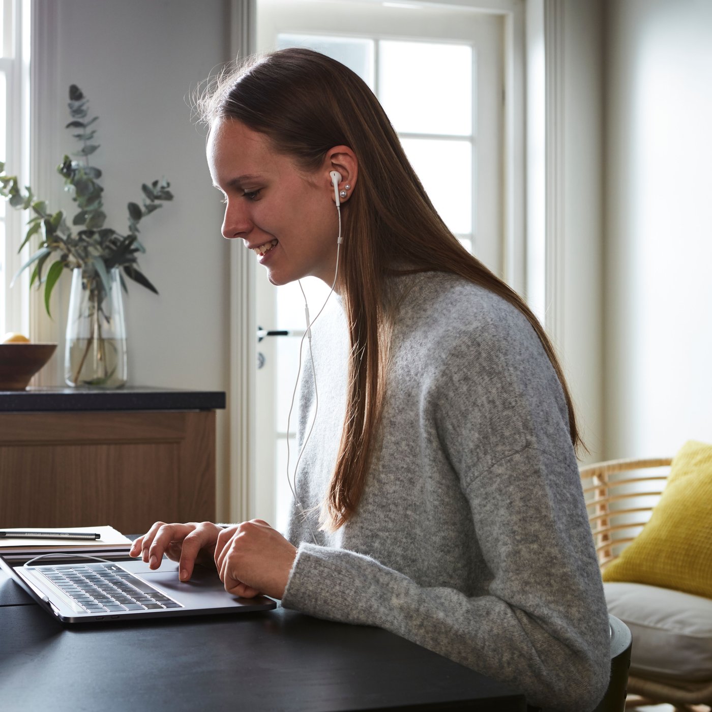 Une personne aux cheveux longs et vêtue d'un pull gris est assise à une table et travaille sur un ordinateur portable. Elle porte des écouteurs.