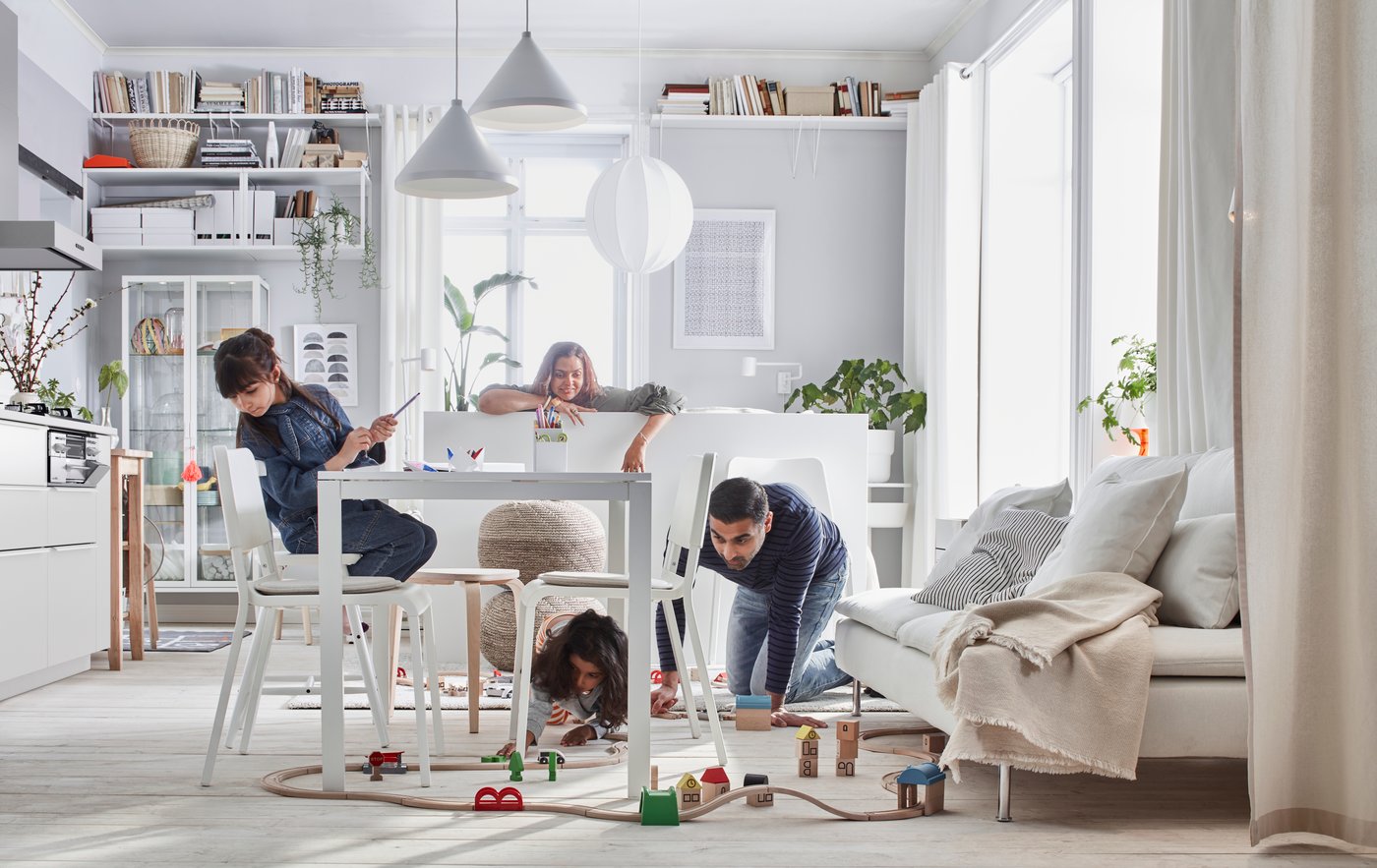 Familie in einem Wohnzimmer mit einer MELLTORP TEODORES Essgruppe neben einem SÖDERHAMN Sofa, unter der ein Mädchen mit einer LILLABO Eisenbahn spielt.