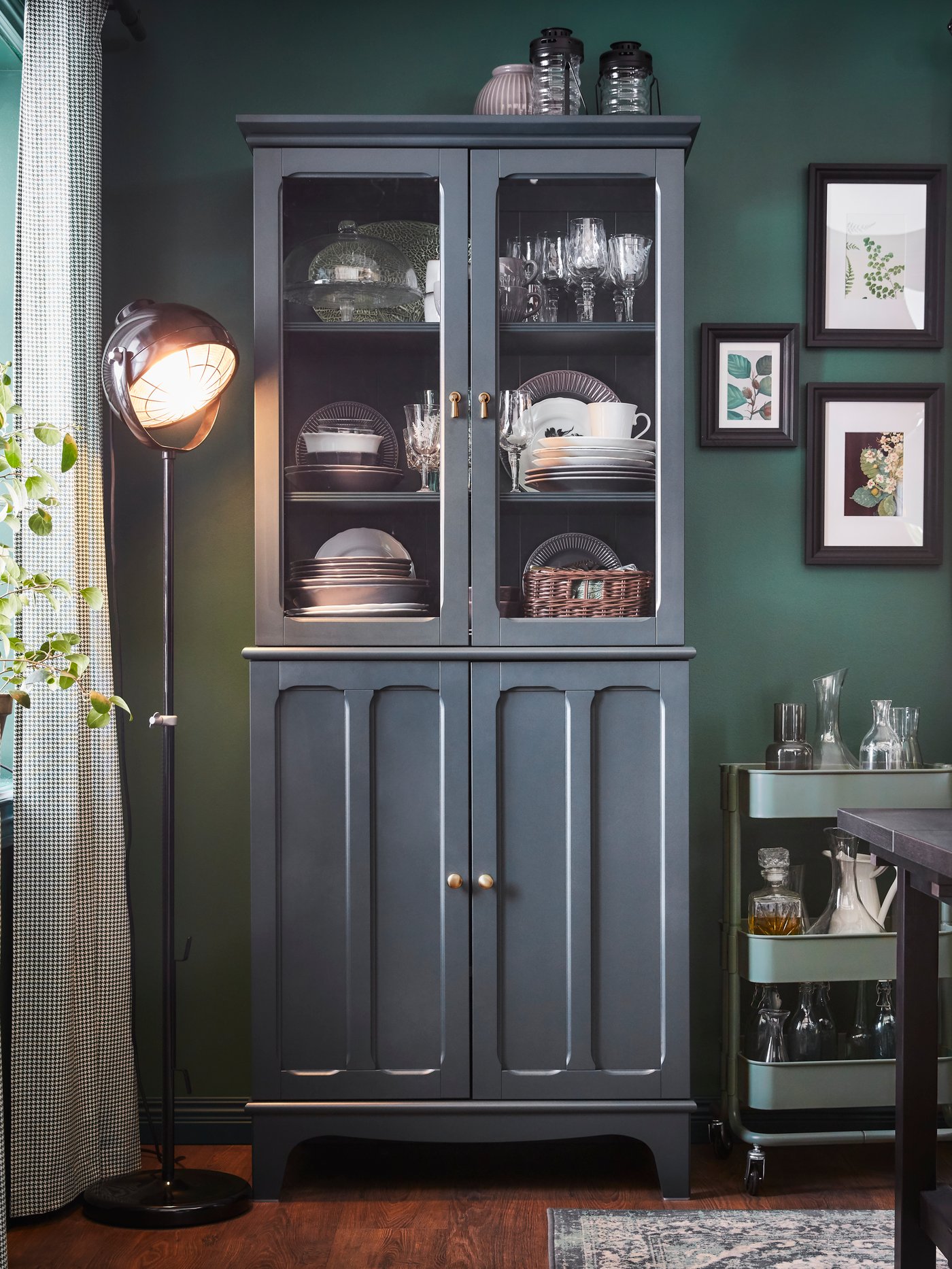 Dinnerware is stored in a blue-green cabinet with glass doors. A black floor lamp and a grey-green trolley stand beside it.