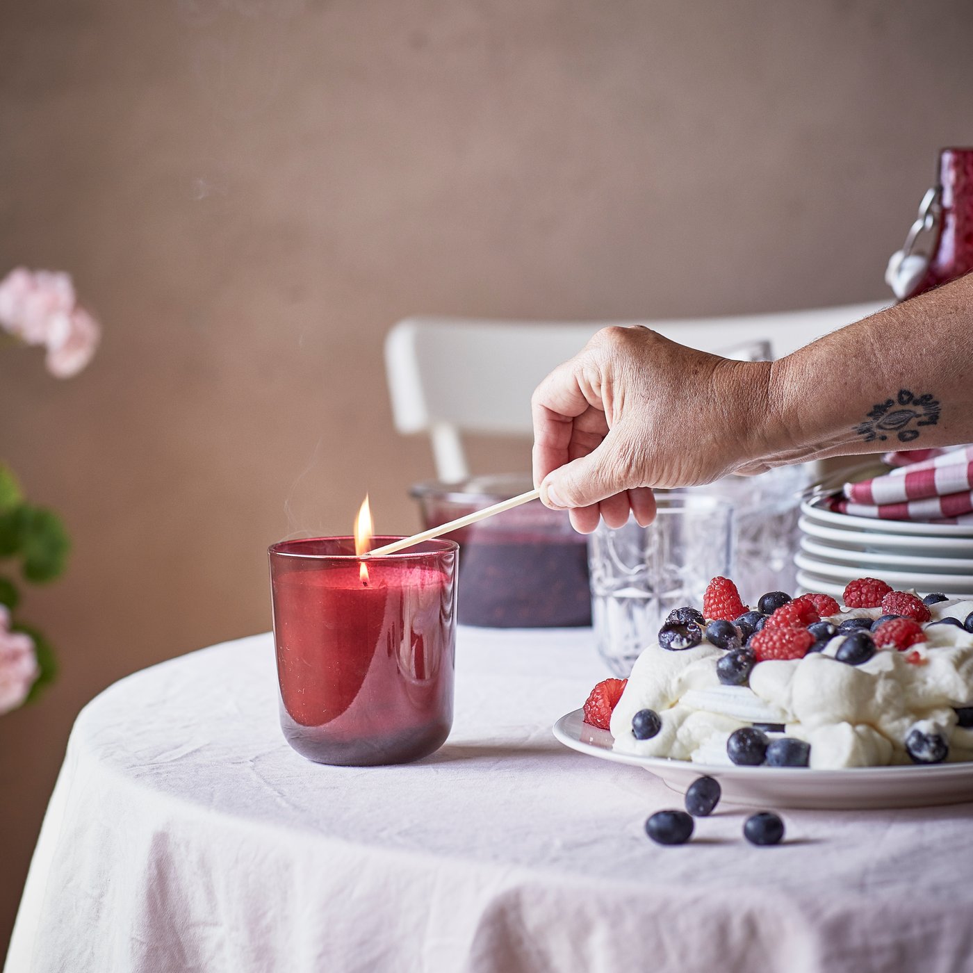 Una tavola con una tovaglia bianca è appoggiata una candela rossa in un contenitore in vetro con una persona che sta accendendo la miccia.