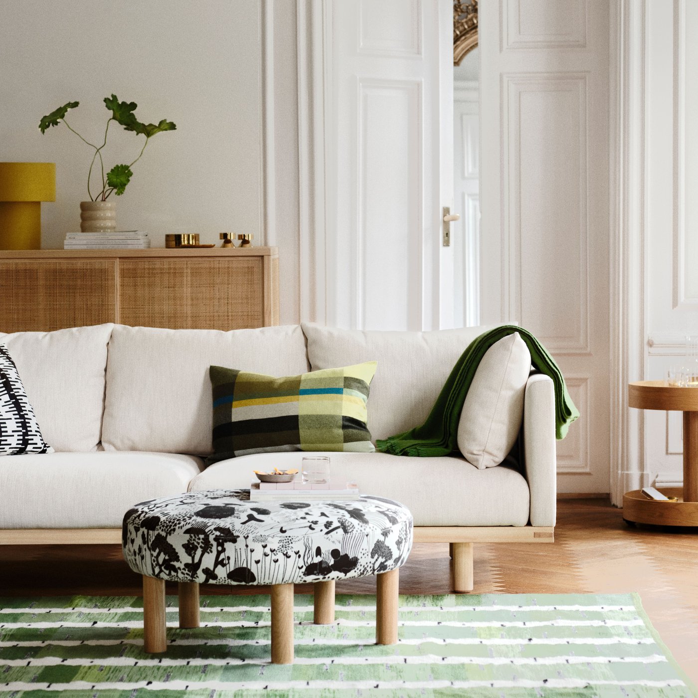 Ein Wohnzimmer mit einem 3-Sitzer-Sofa STOCKHOLM 2025, Kissen und einer Decke darauf und einem schwarz-weißen Pouf auf einem Teppich davor.