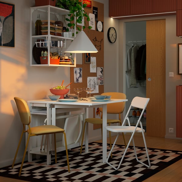 A VIHALS gateleg table with chairs around it is standing in a small dining area and there are things stored on a shelf above.