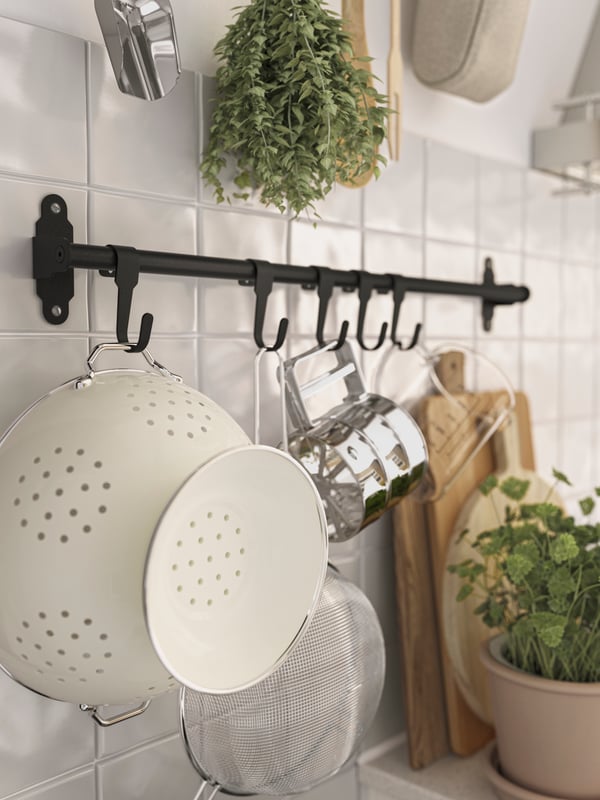 Close-up of black metal wall rail with hooks hung with white enamel colander and glass measuring jug, with potted herbs and wood chopping boards nearby.