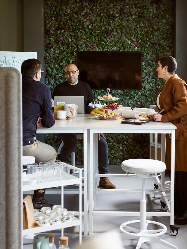 A wall covered in FEJKA plants with a TV screen in the middle is in a meeting area, where people are gathered at high tables.