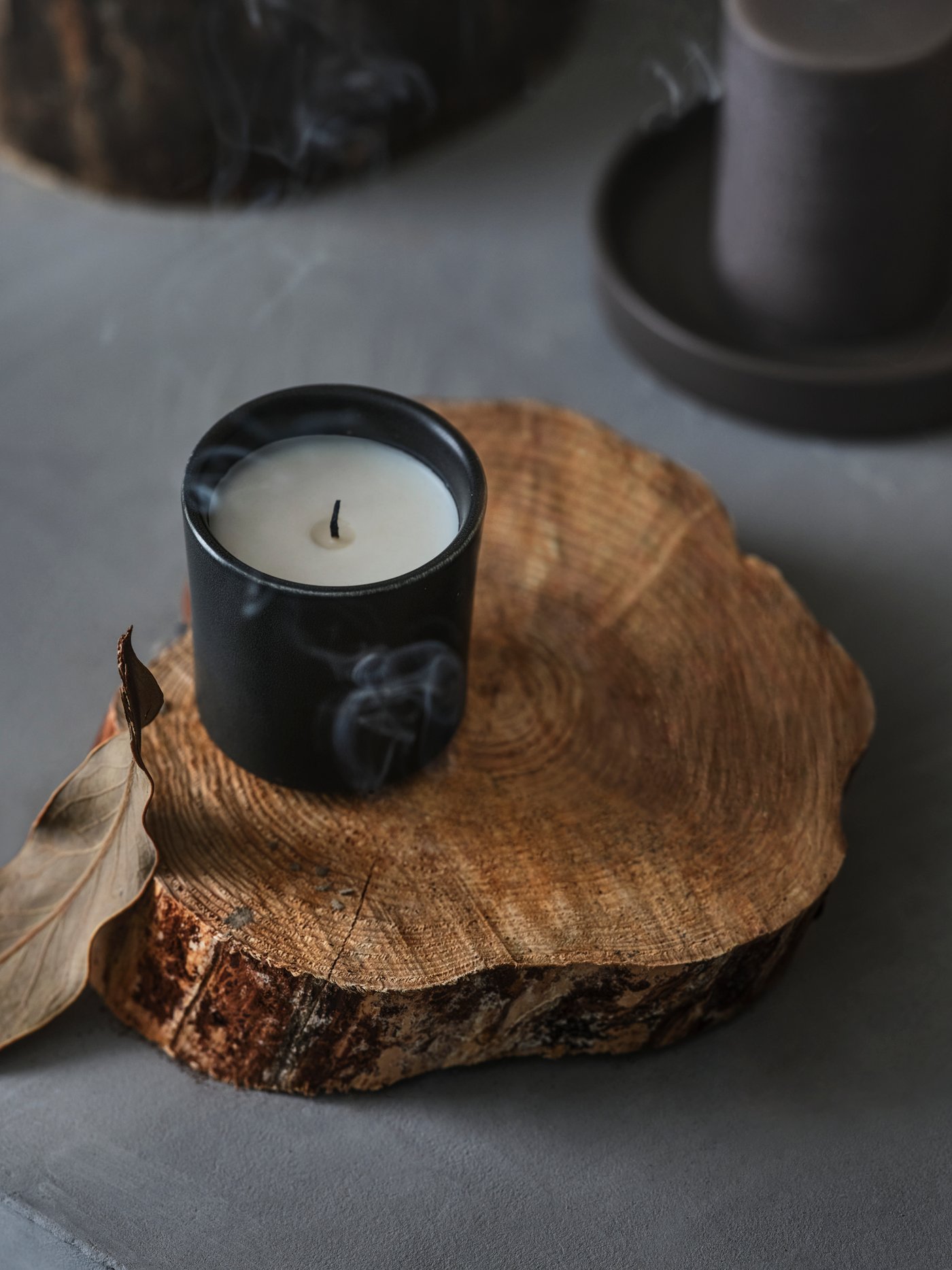 Une bougie parfumée BEHJÄRTAD dans un pot en céramique se trouve sur un morceau de bois et un nuage de fumée s'en détache.
