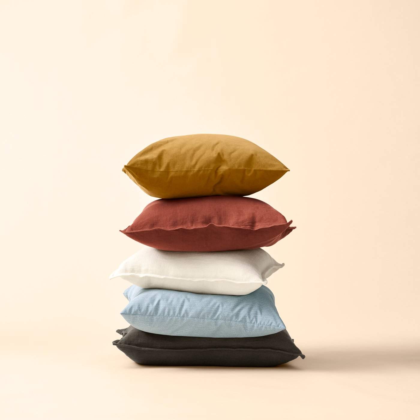 Five stacked cushions, one of them with a 50-centimetre, yellow-brown SANELA cushion cover, against a pale-yellow background.