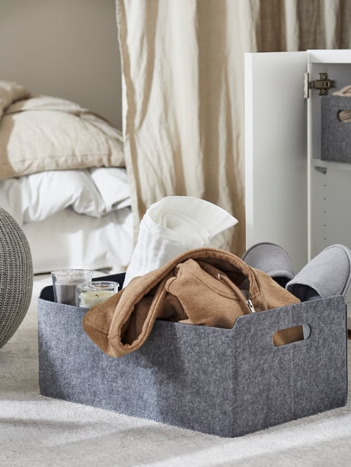 Close up on living room floor with open storage unit and grey fabric storage box filled with throw, slippers, and candles.