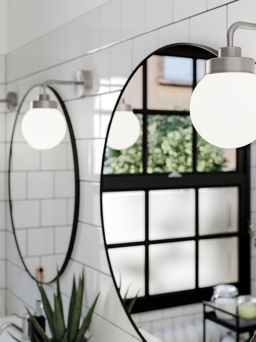Two round LINDBYN mirrors side by side on a white-tiled wall, mirroring a window with greenery outside, plus wall lamps.