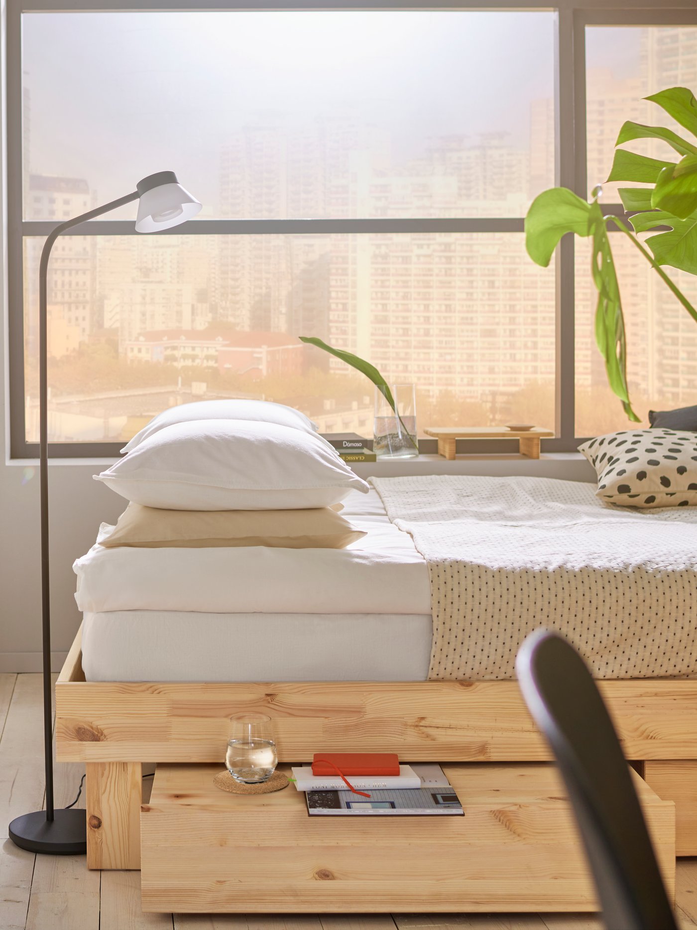 Spálňa v neutrálnom štýle s veľkým oknom, posteľ z neošetreného dreva so zásuvkou pod ňou, béžová posteľná bielizeň.