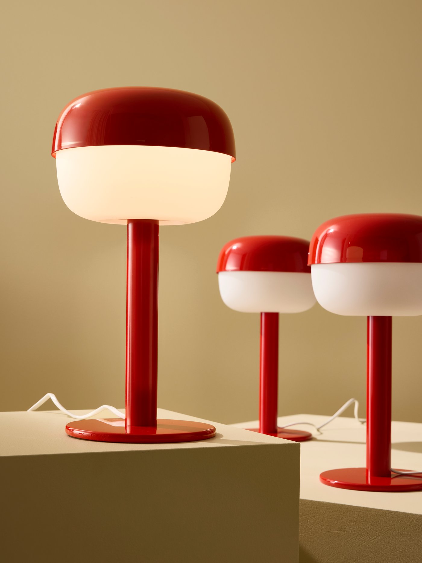 Tre bordslampor i retrostil, röda och vita, placerade på socklar mot en neutral bakgrund.