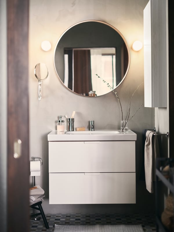 A round mirror above a GODMORGON/ODENSVIK wash-stand with two wall lamps on either side. Toiletries are on the basin.