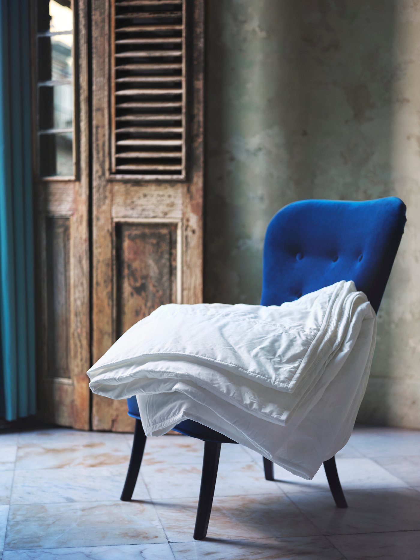 A bright blue velvet chair with a white duvet draped over the seat, set against a rustic wooden door on a tiled surface.