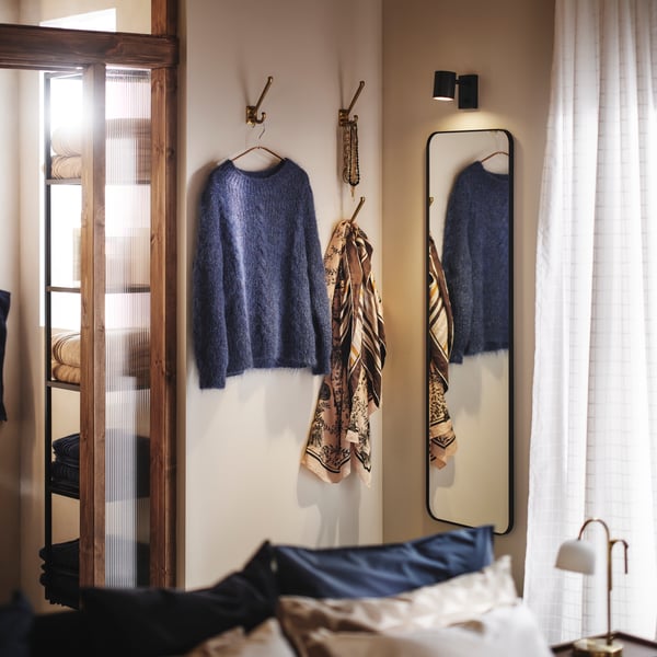 Corner of a bedroom with LINDBYN mirror and hooks for clothes and accessories on the walls. Glass doors and window at the sides.