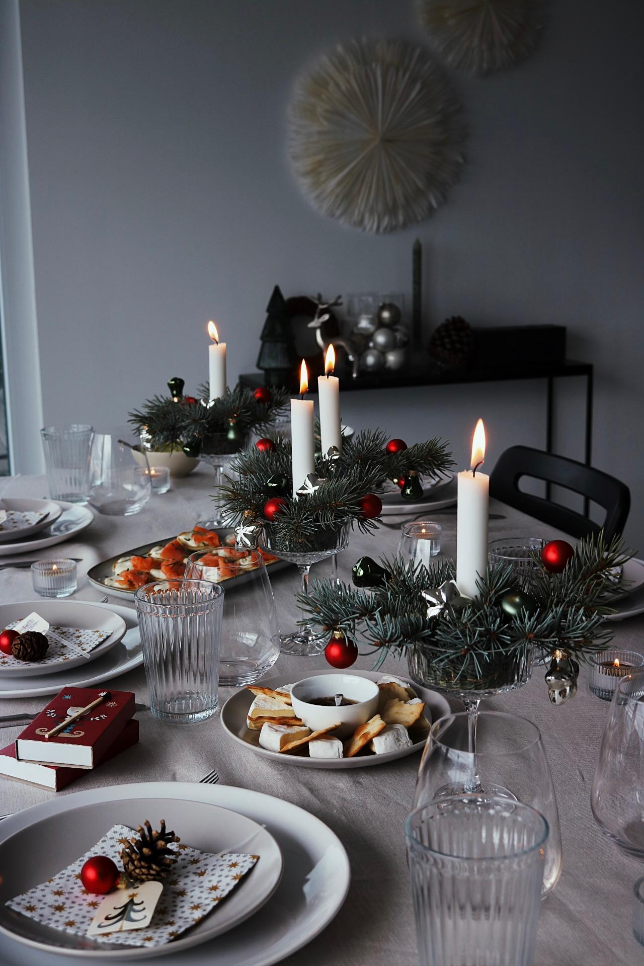 Festlich gedeckter Esstisch mit brennenden weissen Kerzen, immergrünen Tischdekorationen, roten Ornamenten und einer eleganten Tischdekoration. Gemütliche und einladende Weihnachtsstimmung.