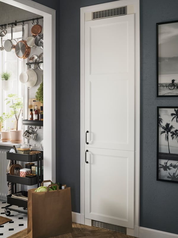 A fridge/freezer with matte white AXSTAD fronts are placed just outside a small kitchen with a black RÅSKOG trolley.