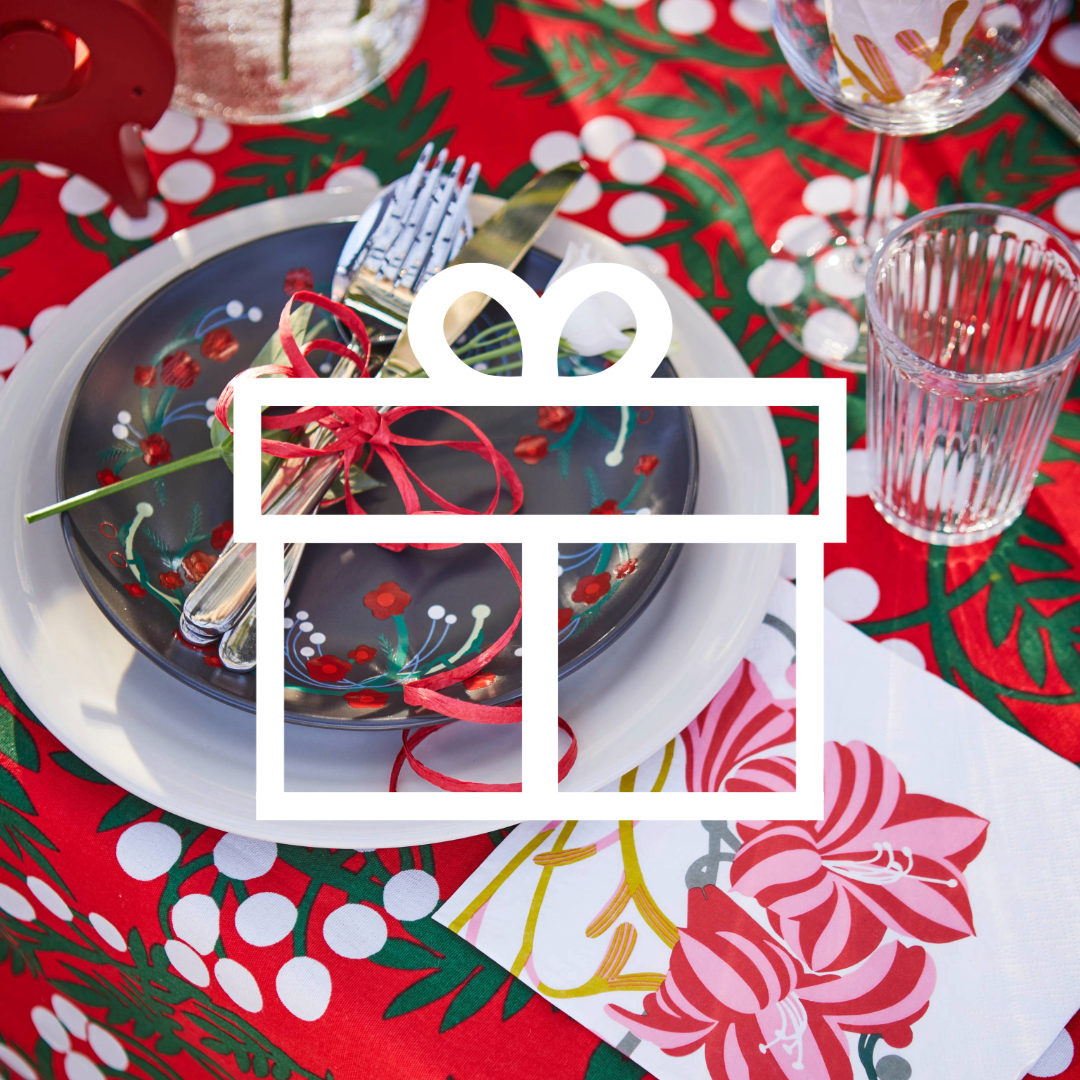 Et dækket bord med service og bestik ovenpå en farverig juledug øverst på billedet er et hvidt ikon af en indpakket gave.