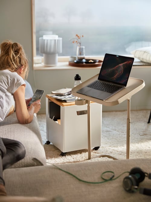 Un salon dans lequel on retrouve une personne allongée sur le canapé, un ordinateur portable placé sur un support pour ordinateur portable, un rangement, un tapis et des housses de coussin.