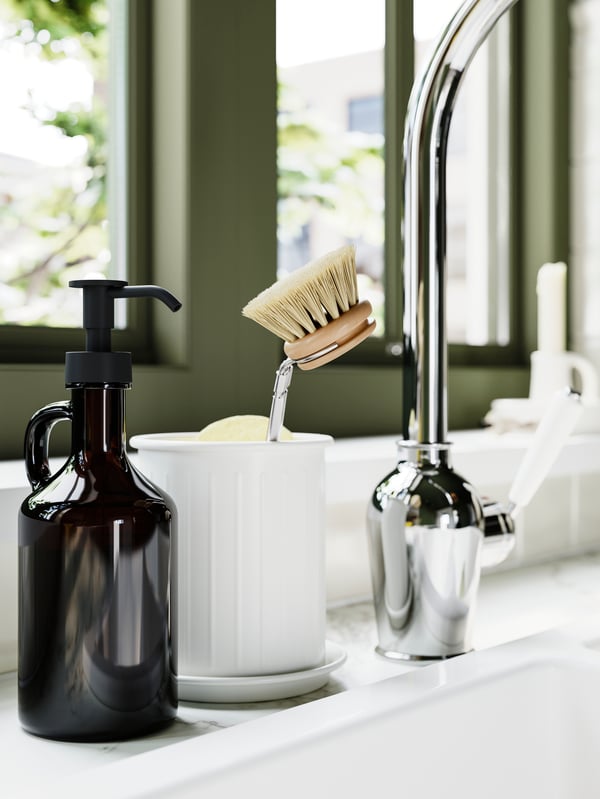 A kitchen sink with a chrome tap, VÄLVÅRDAD cutlery stand holding a dish brush, and a soap dispenser by the window.