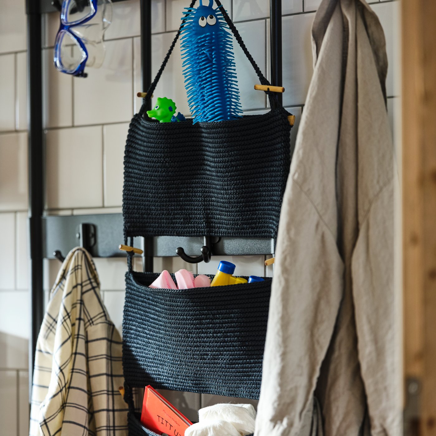 حائط حمام مكسو ببلاط أبيض مع وحدة تخزين معلق NORDRANA باللون الأزرق على علاقة معاطف مع تخزين للأحذية PINNIG باللون الأسود.