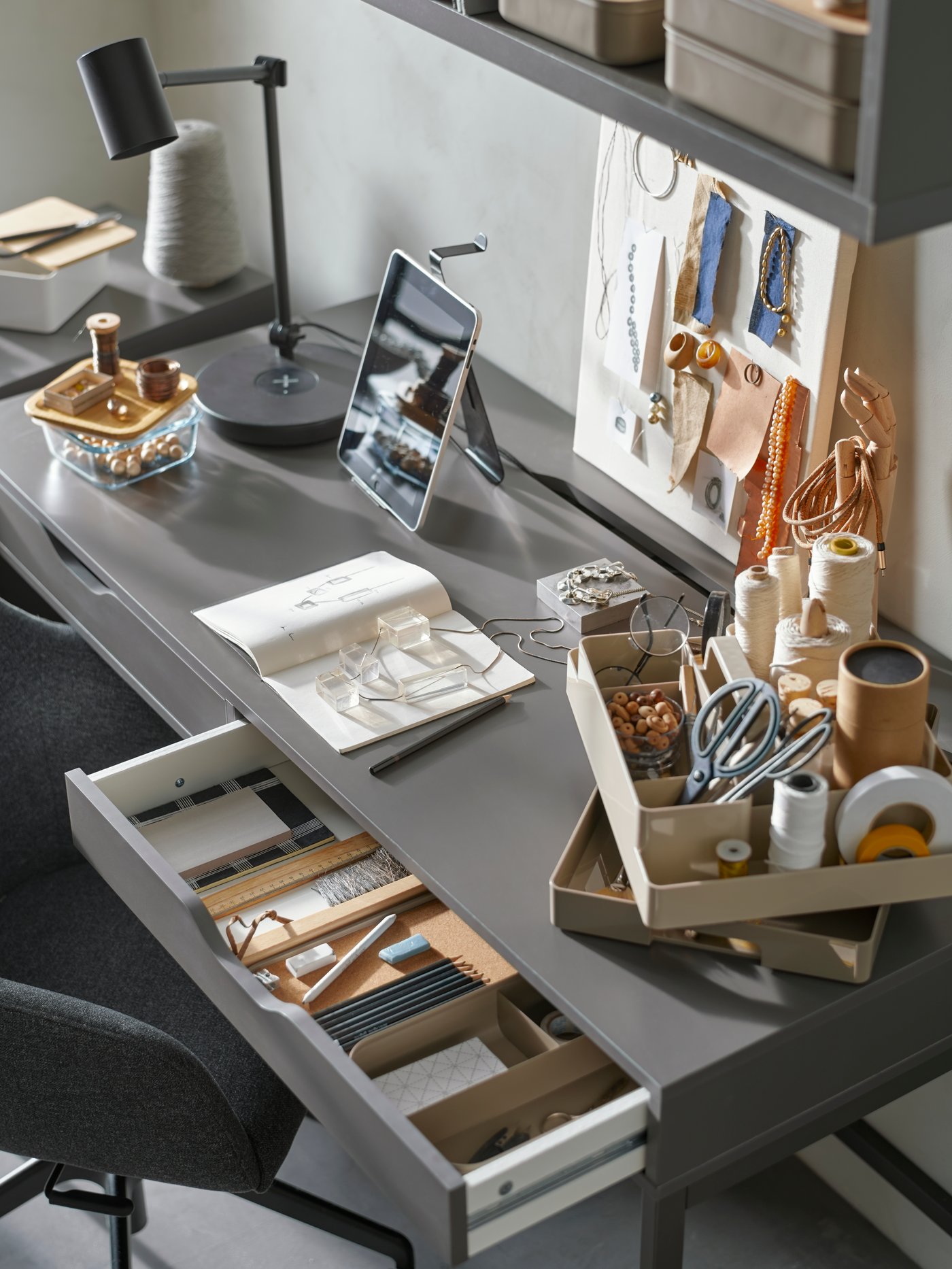 Ein Schreibtisch mit einem Moodboard, einem Skizzenbuch, Materialien zum Basteln von Accessoires, Stiften und einem grau-beigen KUGGIS Schreibtisch-Organizer-Set.