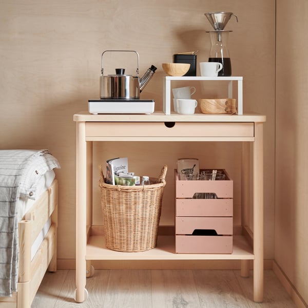 A trolley RÖNNINGE standing next to a bed. There is a portable induction hub TILLREDA with a kettle METTALISK on. In the bottom part of a trolley, there is KAKUSFIKON plant pot next to two stacked KNAGGLIG wooden boxes.