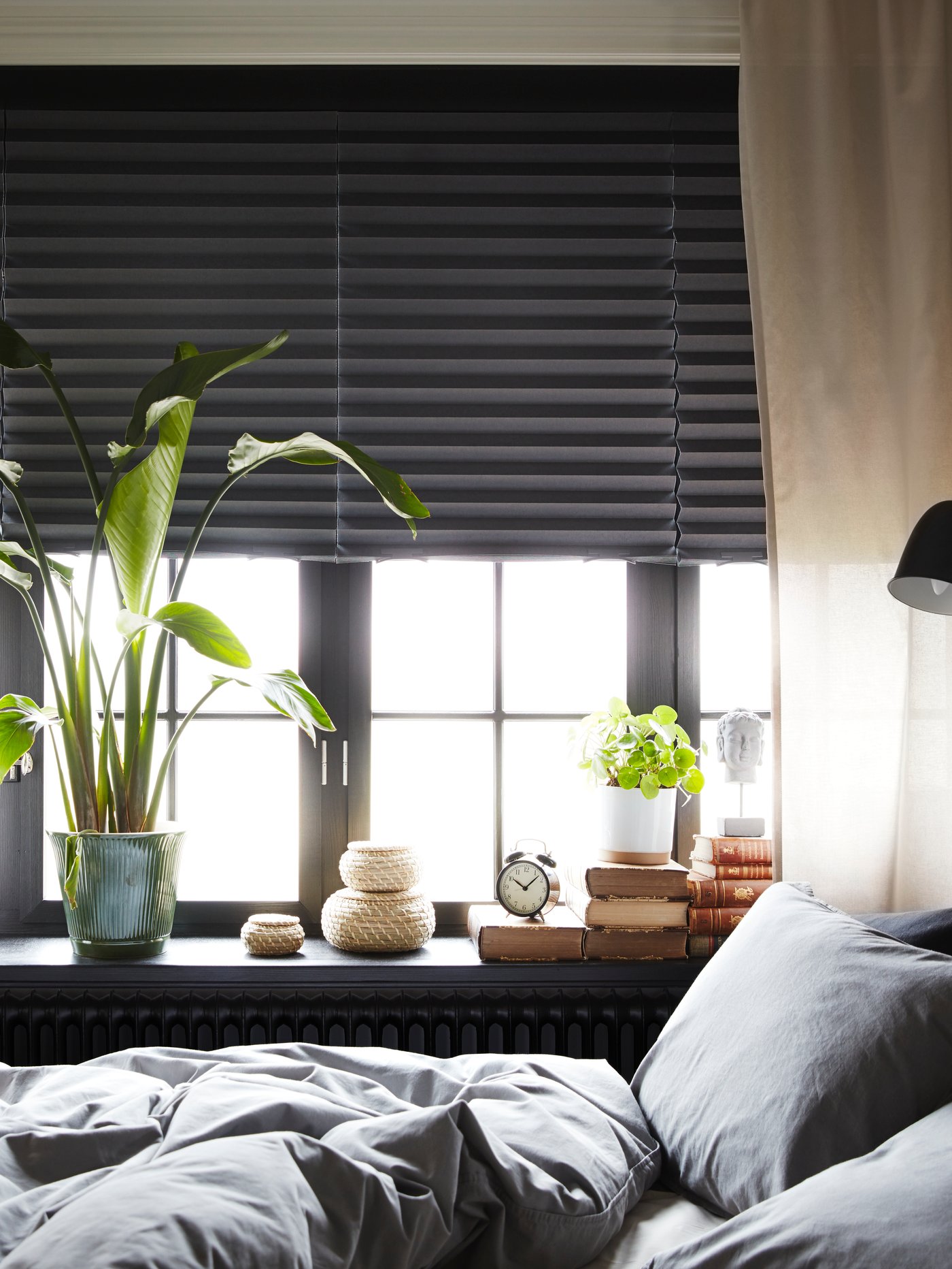 Dormitorio moderno con estores opacos plisados de tela gris junto a macetas y una funda nórdica y dos fundas de almohada grises.