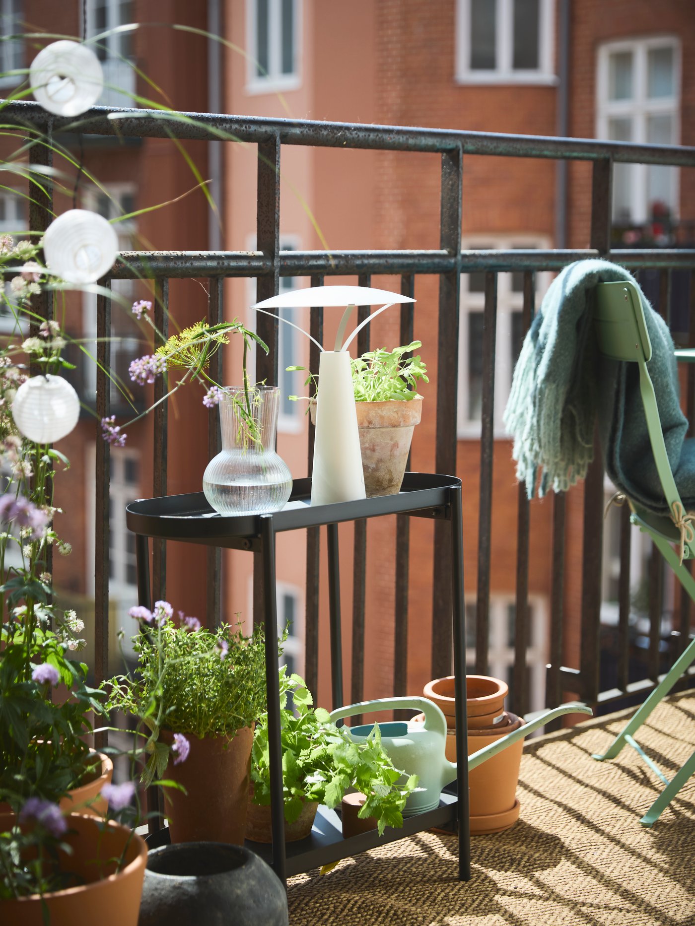 A small balcony in a peaceful setting with green plants, OLIVBLAD plant stand in black, and SOLVINDEN lamp.