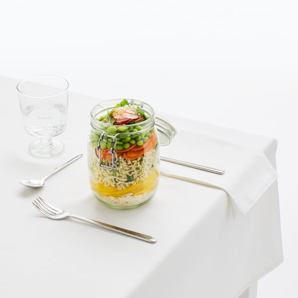 A KORKEN glass jar on a table, containing layers of food: dessert at the bottom, main in the middle and starter on top.