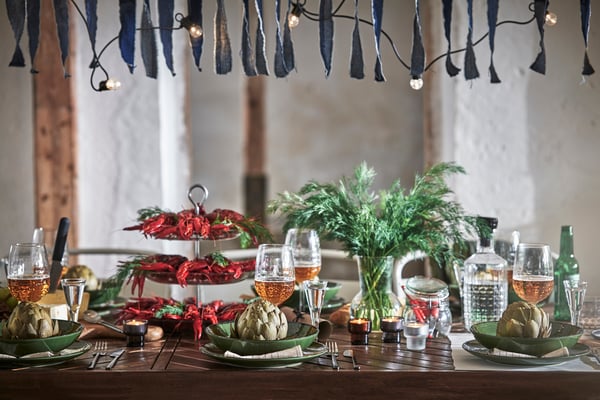 Dukat bord i rustik miljö med kräftor i etagère, kronärtskockor på tallrikar, glas och gröna växter, under blå girlang och ljusslinga.
