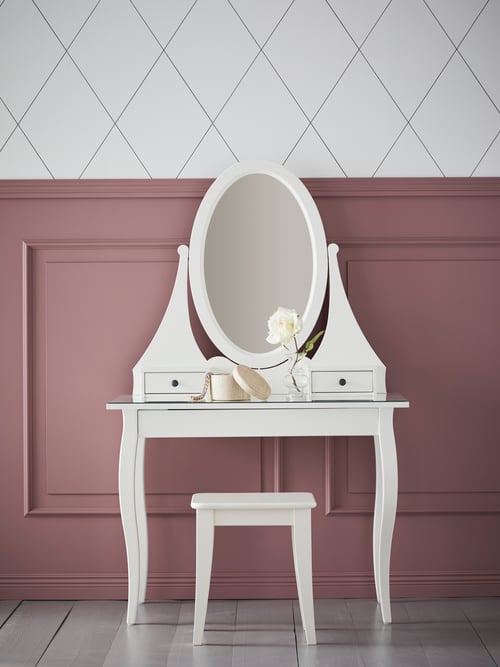 View of room with patterned wall, wooden frame, white dressing table, white stool, box with lid, clear vase, white flower. 