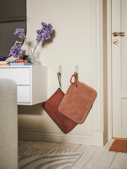 Wohnzimmer mit mehreren KLYKET klappbaren Haken, an der zwei Ledermappen befestigt sind. Daneben ist ein EKET Schrank mit zwei Schubladen zu sehen. Vor der Tür liegt eine TRAMPA Fußmatte.