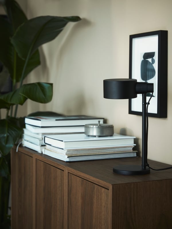 Černá pracovní lampa LÖVMÅNAD a dva komínky knih stojí na hnědém úložném řešení se vzorem ořechu EKET.