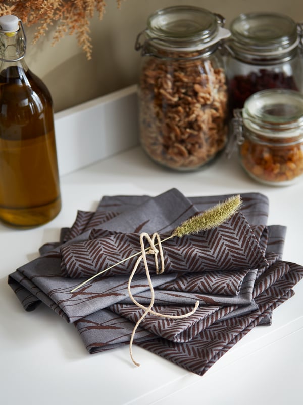 A stack of folded grey HÖSTKVÄLL napkins sit on a sideboard. One napkin is tied with twine and decorated with a dried flower.