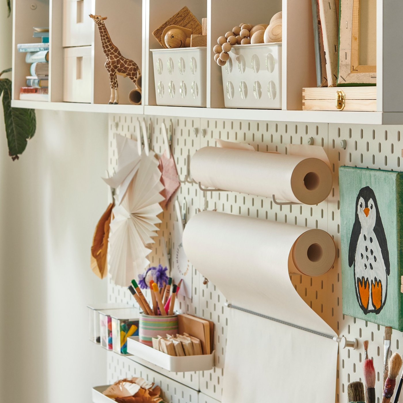 Biela polica s hračkami a krabicami, zavesená nad perforovaným panelom SKÅDIS, na ktorom sú rolky papiera a perá.