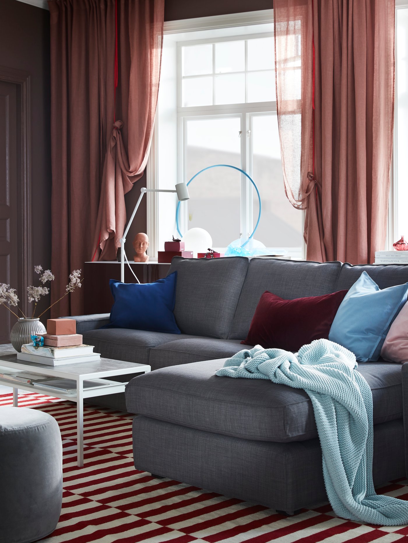 A living room with a dark grey KIVIK sofa with chaise longue and pink LEJONGAP curtains in front of the window.